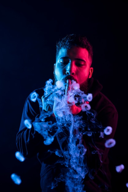 Giovane barbuto uomo arabo che fuma vape e fa anelli di fumo con luci colorate.