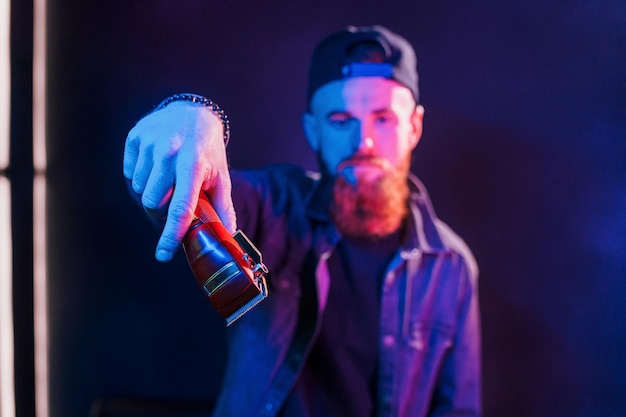 Young bearded barber in cap standing in the studio with neon lighting