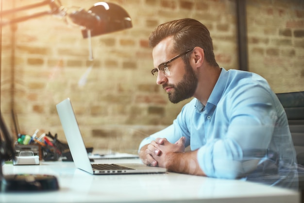 Young bearded businessman in eyeglasses and formal wear looking at laptop