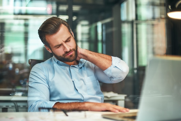 Young bearded businessman in formal wear massaging his neck while sitting in the modern office