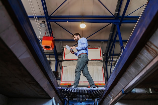 Young bearded businessman holding tablet and walking in garage of shipping firm.