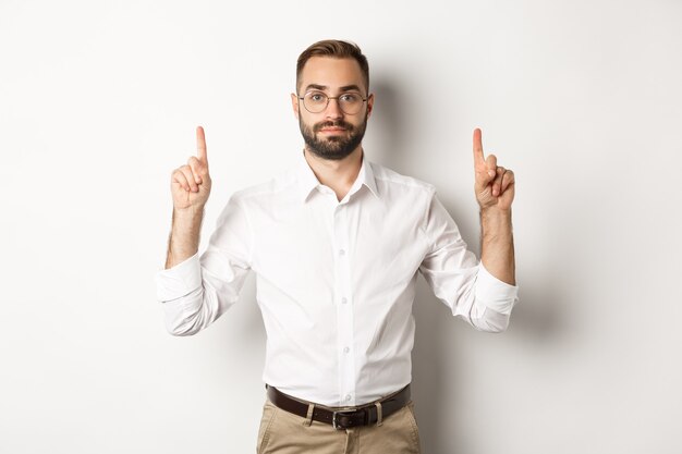 Young bearded businessman pointing fingers up, showing promo offer, standing over white background