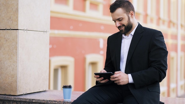 Молодой бородатый бизнесмен читает новости на планшетном компьютере и пьет кофе