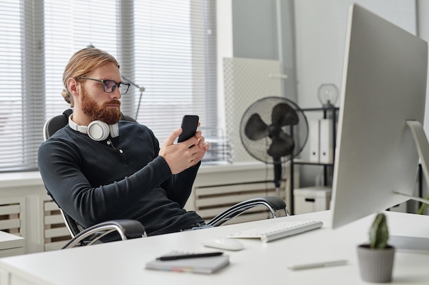 Giovane uomo d'affari barbuto che scorre nello smartphone mentre è seduto sul posto di lavoro