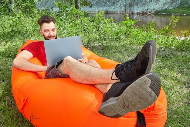 Young bearded businessman with notebook is relaxing on orange inflatable lounger during eco-tourism near river.