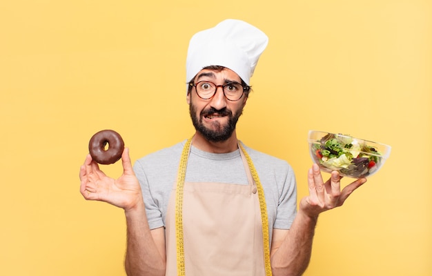 Young bearded chef man with doubting or uncertain expression holding a salad and donut