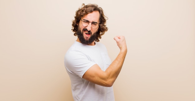 Young bearded crazy man feeling happy, satisfied and powerful, flexing fit and muscular biceps, looking strong after the gym against flat color wall