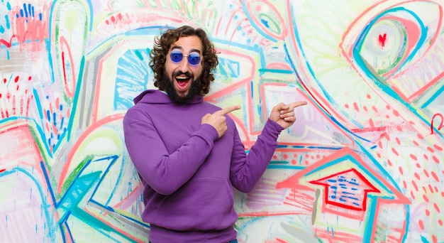 Young bearded crazy man feeling joyful and surprised, smiling with a shocked expression and pointing to the side on graffiti wall