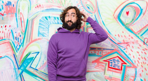 Young bearded crazy man feeling puzzled and confused, scratching head and looking to the side against graffiti wall