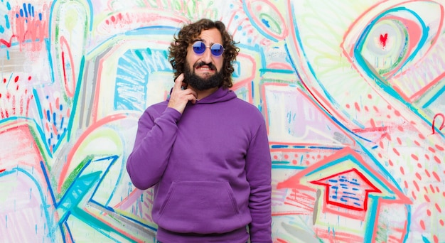 Young bearded crazy man feeling stressed, frustrated and tired, rubbing painful neck, with a worried, troubled look against graffiti wall