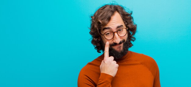 Young bearded crazy man keeping an eye on you, not trusting, watching and staying alert and vigilant against flat color wall