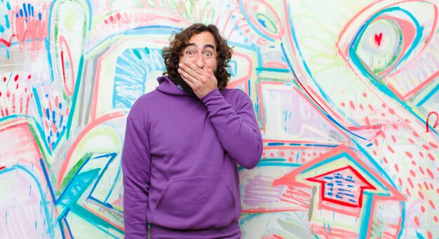 Young bearded crazy man looking happy, cheerful, lucky and surprised covering mouth with both hands on graffiti wall