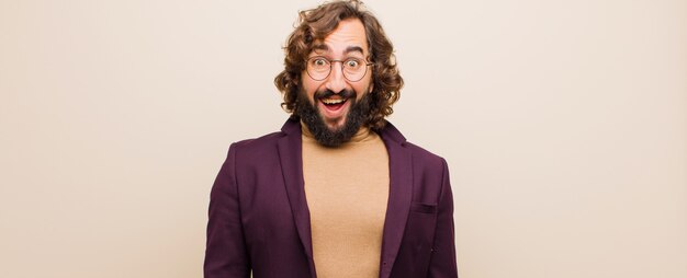 Young bearded crazy man looking happy and pleasantly surprised, excited with a fascinated and shocked expression against flat color wall