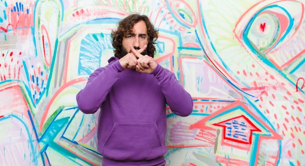 Young bearded crazy man looking serious and displeased with both fingers crossed up front in rejection, asking for silence against graffiti wall