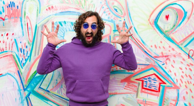 Young bearded crazy man screaming in panic or anger, shocked, terrified or furious, with hands next to head on graffiti wall