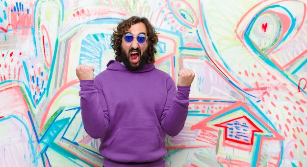 Young bearded crazy man shouting aggressively with an angry expression or with fists clenched celebrating success against graffiti wall