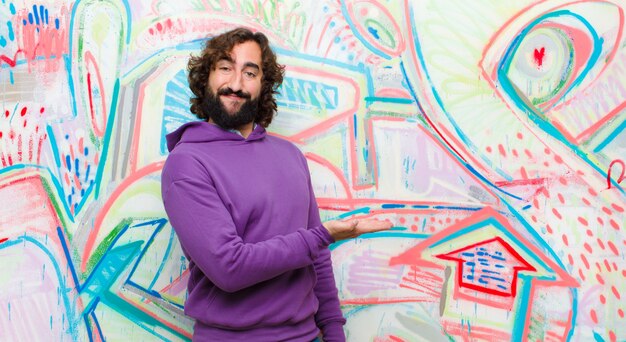 Young bearded crazy man smiling cheerfully, feeling happy and showing a concept in copy space with palm of hand against graffiti wall