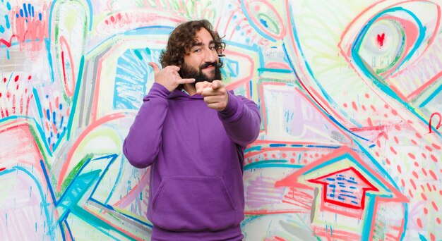 Young bearded crazy man smiling cheerfully and pointing to camera while making a call you later gesture, talking on phone on graffiti wall