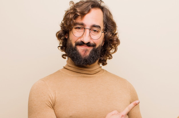 Young bearded crazy man smiling happily and pointing to side and upwards with both hands showing object in copy space