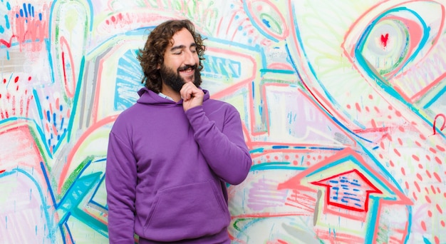 Young bearded crazy man smiling with a happy, confident expression with hand on chin, wondering and looking to the side against graffiti 