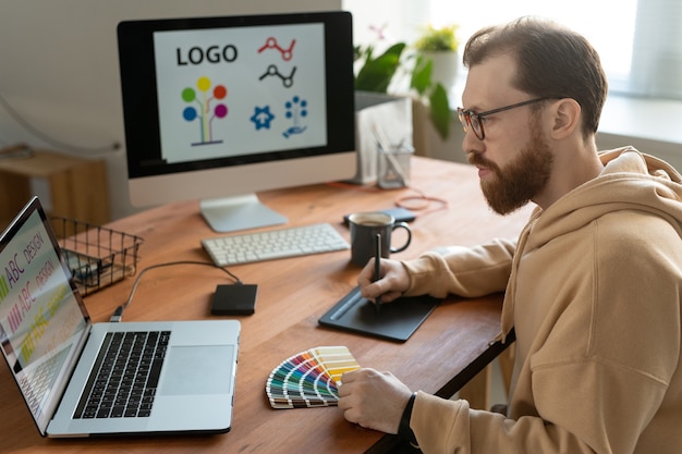 Фото Молодой бородатый креативный дизайнер-фрилансер в очках и повседневной одежде, использующий тачпад, стилус, ноутбук и цветовую палитру