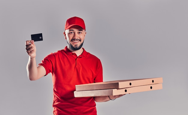 Young bearded delivery man in red uniform with pizza and credit card isolated on gray background.