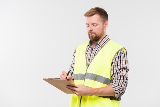 Young bearded engineer with clipboard and pen making working notes while standing in isolation in studio