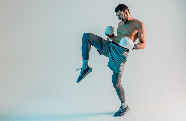 Young bearded european boxer is angry and screaming. Sportsman wear boxing gloves. Isolated on gray background with turquoise light. Studio shoot. Copy space