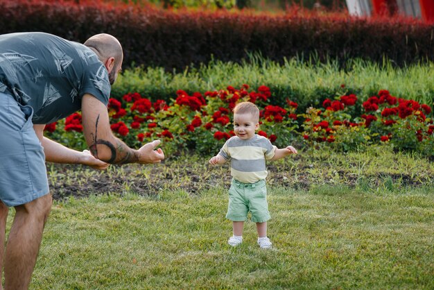 A young bearded father helps and teaches his young son to take his first steps during sunset in the park on the grass.