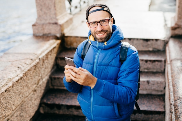 Young bearded guy in black cap and blue anorak holding rucksack standing outdoors with cell phone