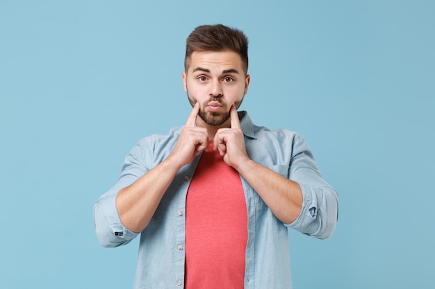 Young bearded guy in casual shirt posing isolated on pastel blue background studio portrait. People sincere emotions lifestyle concept. Mock up copy space. Pointing index fingers on blowing cheeks.