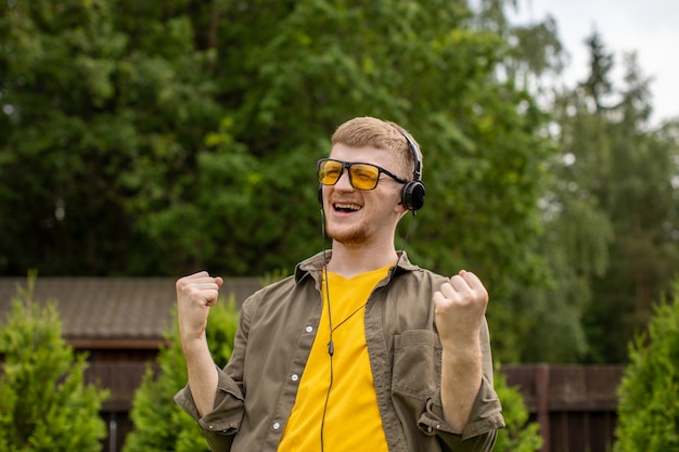 Young bearded guy in yellow glasses dressed casually listening to music in headphones and joyfully singing gesticulating with his hands on green backdrop.