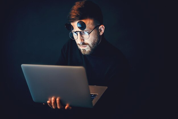 Young bearded hipster man wearing glasses while using laptop over black studio background