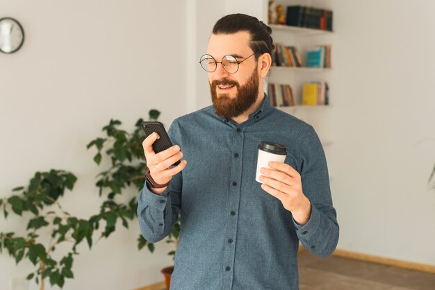 オフィスで若いひげを生やしたヒップスターがコーヒーを飲み、彼の電話で話している