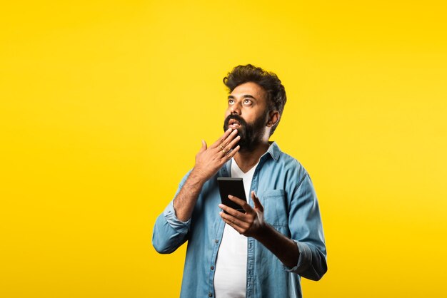 Young bearded Indian man using smartphone, smiling while calling or chatting with friend, standing on yellow