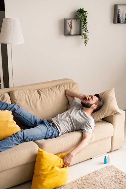 Молодой бородатый мужчина находится в депрессии из-за изоляции коронавируса, лежа на диване и закрывая лицо рукой