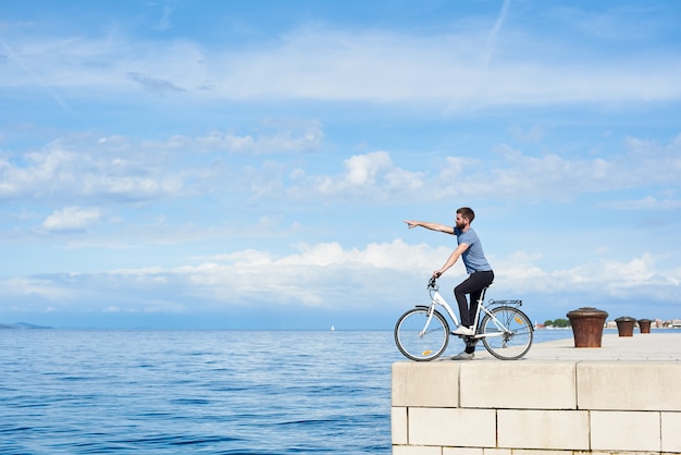 Foto giovane uomo barbuto in bicicletta sul marciapiede di pietra lastricata che punta alla montagna distante sulla riva opposta sull'acqua di mare blu chiaro e piccola barca a vela sullo sfondo