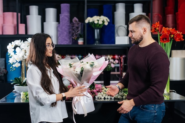 若いひげを生やした男は、居心地の良いフラワーショップで女の子の休日のために美しい花の花束を購入しますフラワーショップでフローリストリーと花束作り中小企業