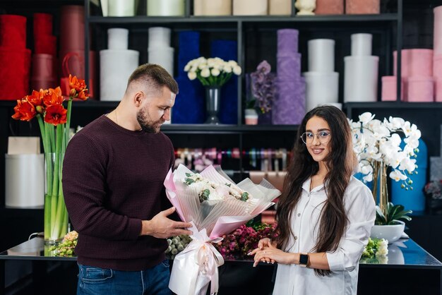 Un giovane uomo barbuto acquista un bellissimo mazzo di fiori per una vacanza di una ragazza in un accogliente negozio di fiori fiorai e bouquet in un negozio di fiori piccola impresa