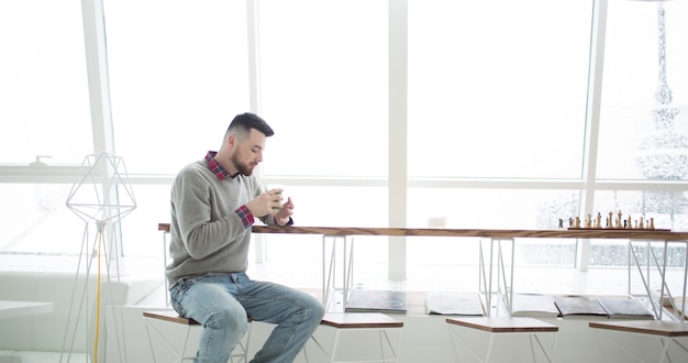 Young bearded man in cafe. he dressed in gray sweater and blue\
jeans. sits on high chair at long table near window.