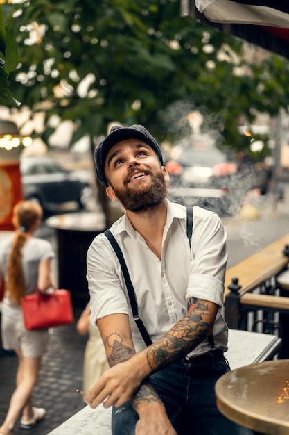 Young bearded man in a cafe on the street smokes a cigarette. Romantic guy in a white shirt cap and suspenders in the city. Peaky Blinders. old fashionable retro.