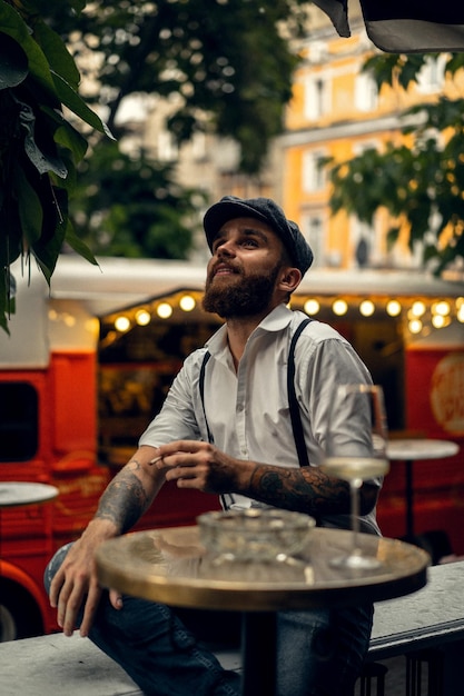 Young bearded man in a cafe on the street smokes a cigarette. Romantic guy in a white shirt cap and suspenders in the city. Peaky Blinders. old fashionable retro.