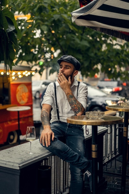 Giovane uomo barbuto in un caffè per strada con un bicchiere di vino. ragazzo romantico con un berretto da camicia bianca e bretelle in città. peaky blinders. vecchia moda retrò.