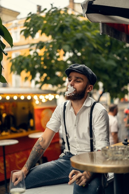 ワインのグラスを持って通りのカフェで若いひげを生やした男。街の白いシャツのキャップとサスペンダーのロマンチックな男。ピーキーブラインダーズ。古いファッションレトロ。