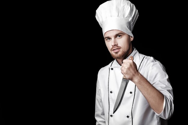 Young bearded man chef in white uniform holds knife on black background