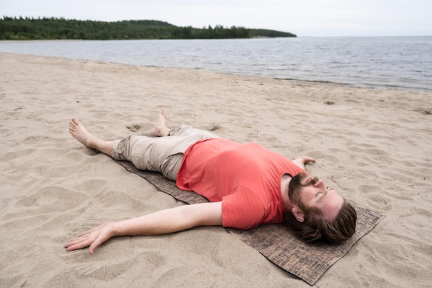 Young bearded man does yoga he does the Savasana exercise relaxes in a Corpse posture lying on a rug