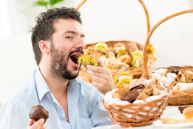 Young bearded man eating a small sandwich,with an expression of enjoyment on his face looking at the camera. In the background you can see woven basket with bakery products.