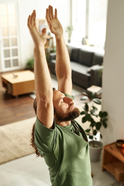 Young bearded man exercising during yoga at home he raising his arms and relaxing with his eyes clos