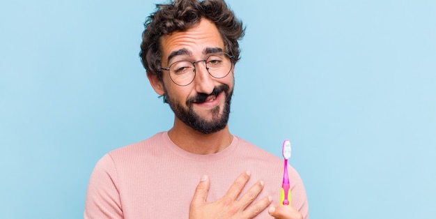 Young bearded man feeling happy and in love, smiling with one hand next to heart and the other stretched up front