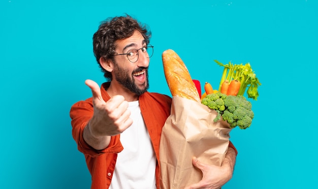 Photo young bearded man feeling proud, carefree, confident and happy, smiling positively with thumbs up and holding a vegetables bag